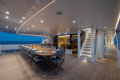 A long, elegant dining table set for a meal on a yacht's deck is in the foreground, with the calm ocean and twilight sky visible in the background.