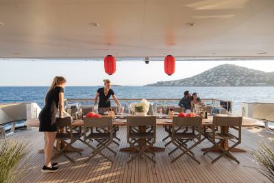 Two women set a long table on a yacht's deck with a couple dining in the background, overlooking a hilly coastline.