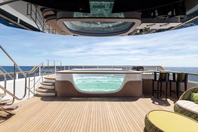 A modern jacuzzi with a glass viewing panel sits on a wooden deck, with the ocean and sky visible in the background.