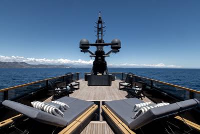 Two plush lounge chairs with striped cushions sit on a wooden deck in the foreground, while a large, dark radar array dominates the background against a clear blue sky and distant coastline.
