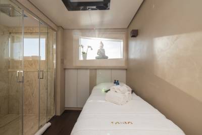 A massage table with towels and bottles sits in the foreground, while a Buddha statue is visible through a window in the background, with a shower stall to the left.