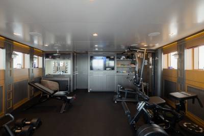 A well-equipped gym with weight benches and machines in the foreground is set against a backdrop of built-in cabinets, a television, and windows.