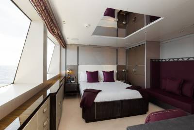 A luxurious yacht bedroom with a large bed and plush seating in the foreground, and a mirrored ceiling and large windows overlooking the ocean in the background.