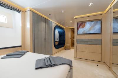 A neatly made bed with gray folded towels in the foreground is set against a modern bedroom with a large closet, a unique oval-shaped seating area, and a television mounted on the wall in the background.