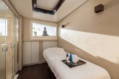 A massage table with towels and bottles sits in the foreground of a spa room, while a Buddha statue is visible through a window in the background.