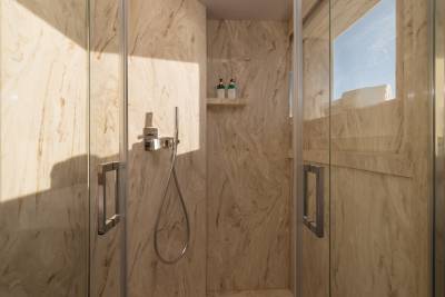 A modern shower stall with marble-patterned walls and glass doors is in the foreground, while a window with a view of a bright sky and building is in the background.