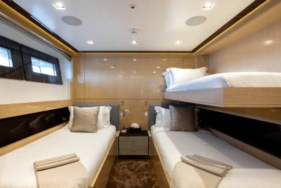 Two twin beds with neatly made white linens and brown accent pillows are in the foreground, while a bunk bed with white linens and a brown accent pillow is in the background, all within a luxuriously appointed cabin.