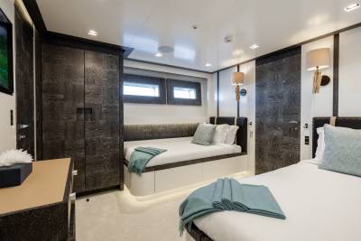 A modern yacht cabin features a built-in bench seat with teal throws in the foreground and dark wood cabinetry and windows in the background.