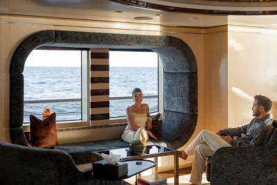 A couple relaxes in a luxurious yacht lounge with a large window overlooking the ocean.