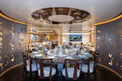 A circular dining table set for a meal is in the foreground, while a luxurious lounge area with sofas and a television is visible in the background.