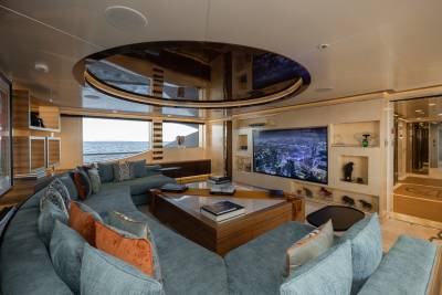 A luxurious yacht lounge with a large sectional sofa and a modern entertainment system is visible, with a view of the ocean through the windows in the background.