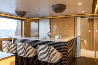 Three striped bar stools are pulled up to a modern, curved bar with drinks on it, while a large window overlooks the ocean in the background.