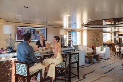 A couple sits at a bar on a yacht, with a bartender serving them, while a lounge area with sofas and tables is visible in the background.