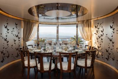 A grand dining table set for a feast is surrounded by walls adorned with butterfly decals, with a panoramic view of rolling hills and a distant city visible through large windows in the background.