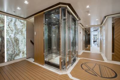A modern glass elevator stands in the foreground of a luxurious hallway with marble accents and wood flooring, leading to a well-lit corridor in the background.