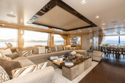 A luxurious yacht lounge with a large sectional sofa and coffee table in the foreground, and a dining area with a table and chairs visible in the background, all overlooking a serene ocean view.