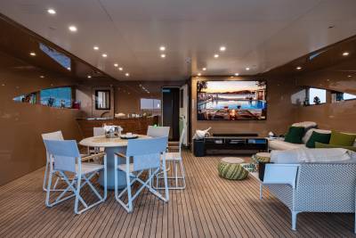 A circular dining table with white chairs sits in the foreground, while a large television displays a scenic view in the background of a luxurious yacht interior.