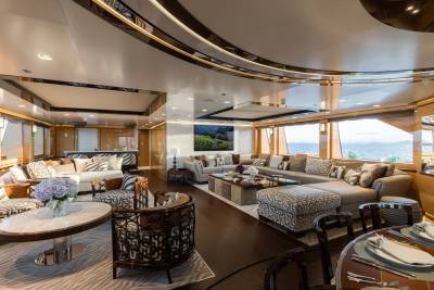 A luxurious yacht interior features a spacious living area with plush seating and a large screen displaying a scenic landscape, while large windows offer a glimpse of the ocean and distant islands.