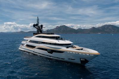 A luxurious white and gold yacht cruises on calm blue water with a mountainous coastline under a cloudy sky in the background.