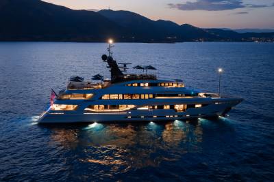 A luxurious yacht with illuminated windows and underwater lights floats on calm, dark water, with a silhouetted coastline and twilight sky in the background.