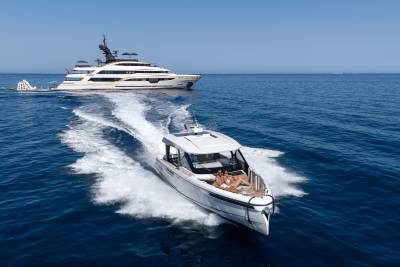 A sleek white speedboat with a person lounging on the bow cuts through the blue ocean, leaving a foamy wake, while a large, luxurious yacht sails in the distance under a clear sky.
