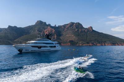 A large white yacht floats on calm blue water in front of rugged, tree-covered mountains, while a small inflatable boat speeds by in the foreground.