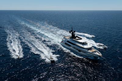 A large white yacht cruises through the deep blue ocean, leaving a frothy wake, while several jet skis race alongside it.