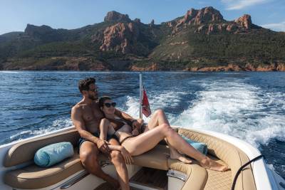 A couple relaxes on the back of a boat with a wake trailing behind them, as rugged, tree-covered mountains rise in the distance.