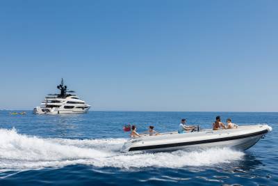 A white inflatable boat with four people speeds across the blue ocean in the foreground, while a large white yacht with black accents sails in the background under a clear sky.