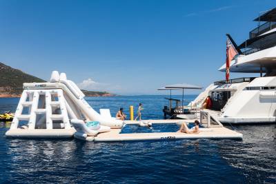 People relax on an inflatable water park and pool attached to a large yacht, with a hilly coastline in the background.