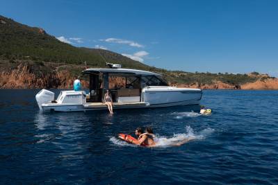 People enjoy a sunny day on the water, with a modern yacht anchored near a lush, mountainous coastline.