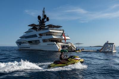 A yellow jet ski speeds across the water in the foreground, with a large white yacht and inflatable water slide anchored in the background under a clear blue sky.