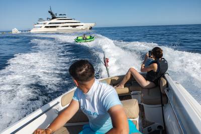 In the foreground, two people are enjoying a ride on a speedboat, while in the background, a large yacht sails on the ocean.