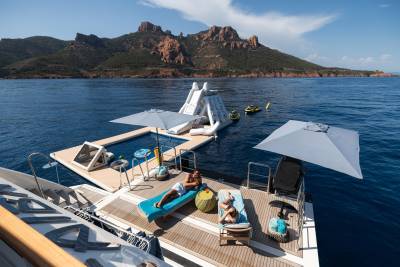 Two people relax on a yacht's deck in the foreground, while a floating water park with slides and jet skis is anchored in the blue sea before a rugged, mountainous coastline in the background.