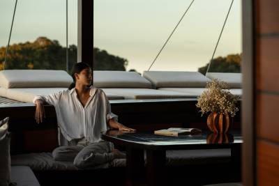 A woman sits on a cushioned bench in the foreground, with a table holding a vase of flowers and a book, while a lush, green landscape is visible in the background.
