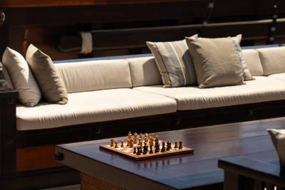 A chess set rests on a dark wooden table in the foreground, with a long, cushioned outdoor sofa and pillows in the background.