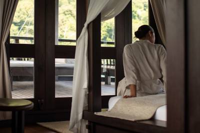 A woman in a robe sits on a bed in the foreground, looking out at a wooden deck and lush greenery through large windows in the background.