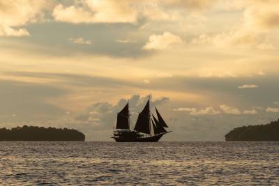 A silhouetted schooner sails on calm water between two islands under a dramatic, cloud-filled sky.