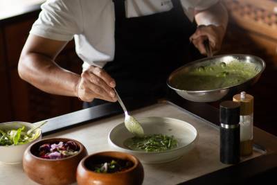 A chef plates a green sauce from a pan into a bowl, with three small bowls of ingredients in the foreground and a salt and pepper grinder to the right.