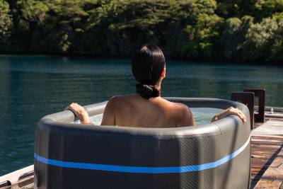 A person relaxes in a hot tub on a wooden deck, with a calm blue body of water and lush green trees in the background.