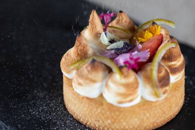 A delicate lemon meringue tart adorned with edible flowers and lime zest sits on a dark surface, with a dusting of sugar falling onto it.