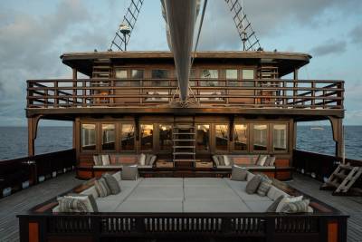 A spacious, cushioned deck with seating in the foreground leads to a multi-level wooden boat with numerous windows and railings, set against a calm ocean and cloudy sky in the background.