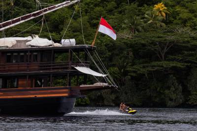 A jet ski speeds past a large wooden boat flying an Indonesian flag, with a lush green jungle in the background.