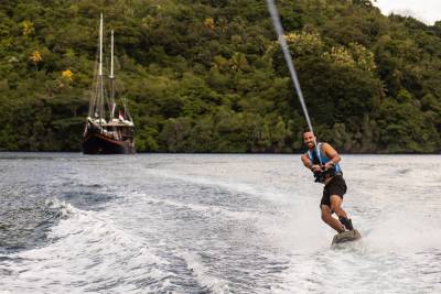 A man wakeboards through choppy water in the foreground, with a large, dark sailing ship and lush green hills in the background.