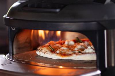 A pizza with melted cheese and red sauce cooks inside a black oven with flames visible in the background.