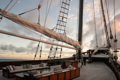 A luxurious wooden deck with plush seating in the foreground is set against a dramatic sky with clouds and the rigging of a sailing ship in the background.