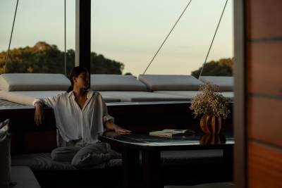 A woman in a white shirt relaxes on a cushioned bench in the foreground, with a table holding a book and flowers, while a row of lounge chairs and a tree-covered hillside are visible in the background.