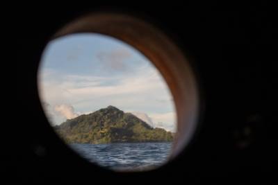 A lush, green island rises from the blue ocean, viewed through a dark, circular porthole.