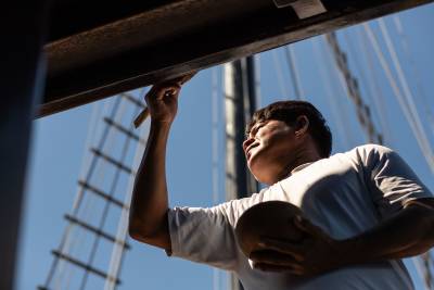 A man in a white shirt paints a wooden beam while holding a round object, with a clear blue sky and rigging visible in the background.