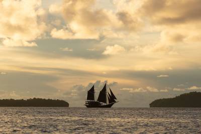 A silhouetted schooner sails on calm water between two tree-covered islands under a dramatic, cloud-filled sky.